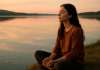 Curso Consciencia – Día 29: La paz está aquí Una mujer sentada en quietud total con los ojos cerrados junto a un lago.